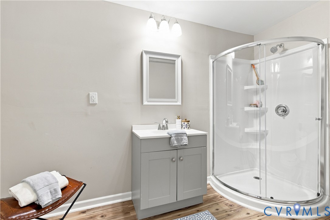 Wood-finish flooring, single vanity with light grey cabinetry, white countertop, framed mirror, and a curved glass shower enclosure with built-in shelving
