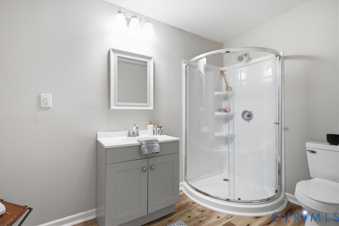 Modern bathroom featuring a curved glass shower enclosure with chrome fixtures, gray vanity cabinetry with integrated sink and countertop, framed mirror with integrated lighting, wood-finish flooring, and white ceramic toilet
