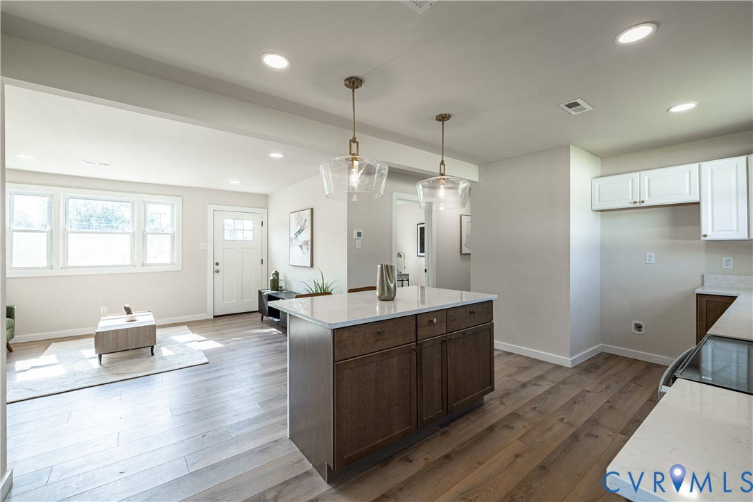 Open-concept living space with wood-finish flooring, recessed lighting, a kitchen island with a light-toned countertop, and white kitchen cabinetry Open-concept living space with wood-finish flooring, recessed lighting, a kitchen island with a light-toned countertop, and white kitchen cabinetry