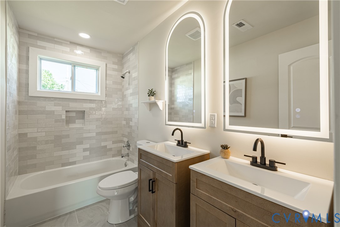 Spa-inspired primary bathroom featuring dual wood-finish vanities with white quartz countertops, matte black faucetry, and illuminated arched mirrors Spa-inspired primary bathroom featuring dual wood-finish vanities with white quartz countertops, matte black faucetry, and illuminated arched mirrors