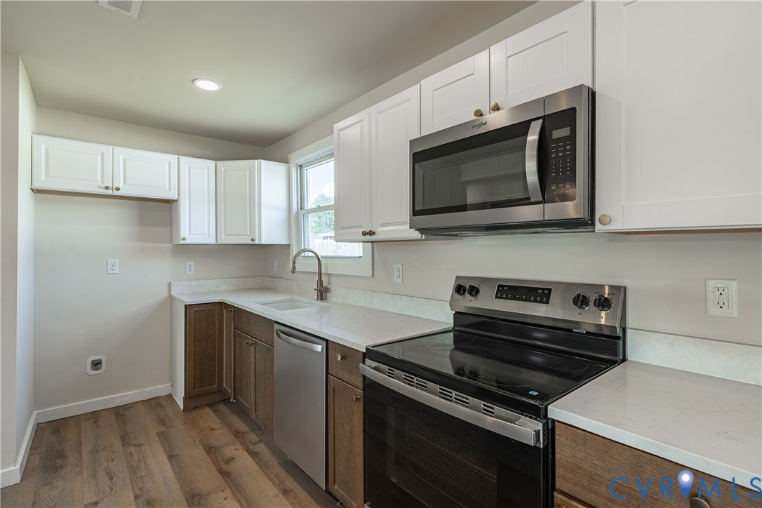 Kitchen featuring white upper cabinetry, wood-finish lower cabinetry, light-toned countertops, stainless steel appliances, and wood-finish flooring Kitchen featuring white upper cabinetry, wood-finish lower cabinetry, light-toned countertops, stainless steel appliances, and wood-finish flooring