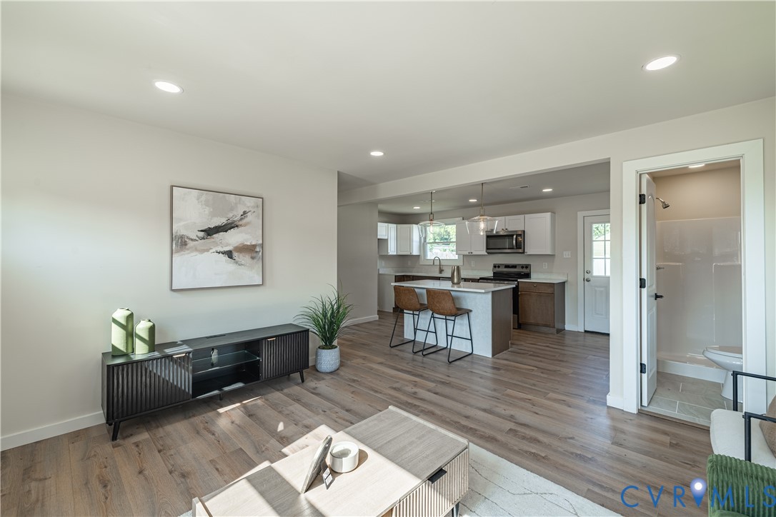Open-concept living space featuring recessed lighting, wood-finish flooring, a kitchen island with seating, white shaker cabinetry, and a full bathroom with a shower Open-concept living space featuring recessed lighting, wood-finish flooring, a kitchen island with seating, white shaker cabinetry, and a full bathroom with a shower