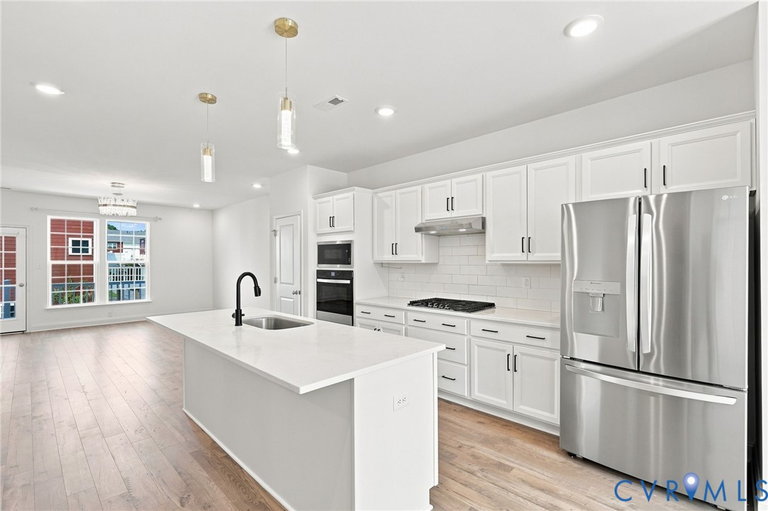 Open-concept kitchen featuring an island with a sink, stainless steel appliances, white cabinetry, and wood-finish flooring