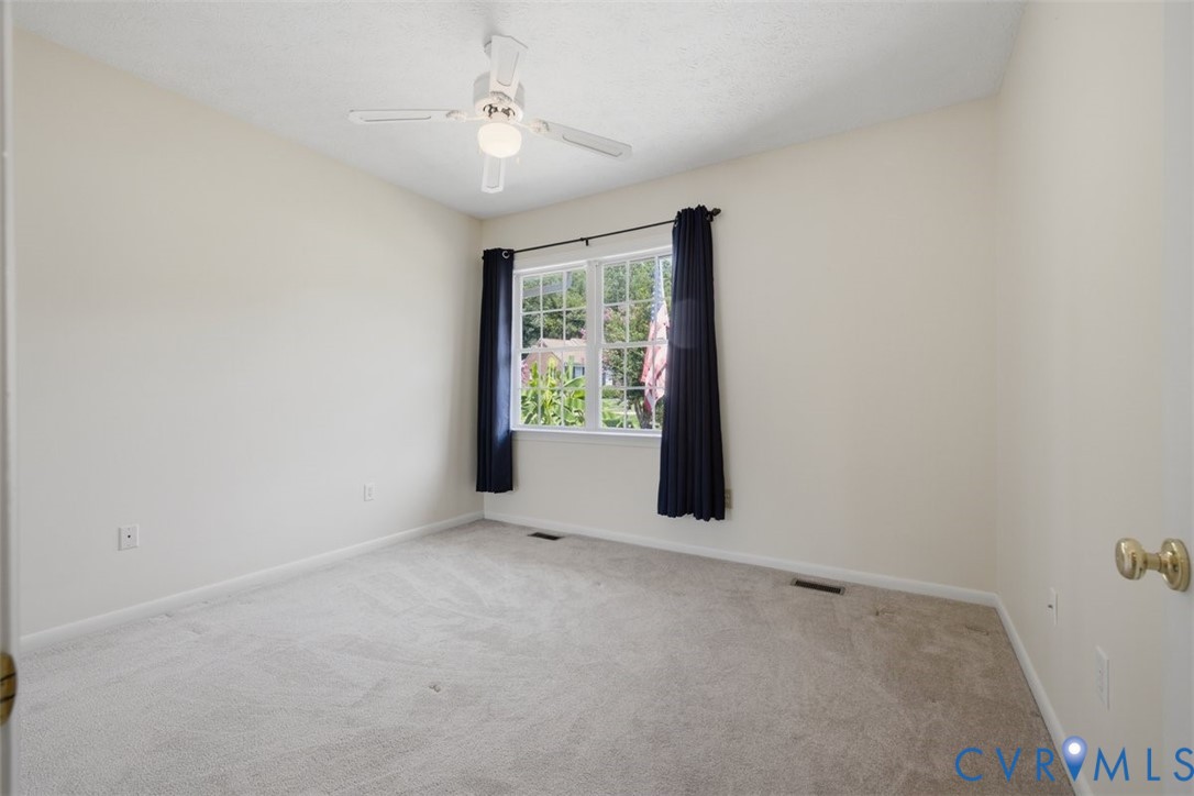 Bright secondary bedroom featuring carpet flooring, a ceiling fan with integrated lighting, and a large double-hung window with black curtain rods framing natural light and outdoor greenery