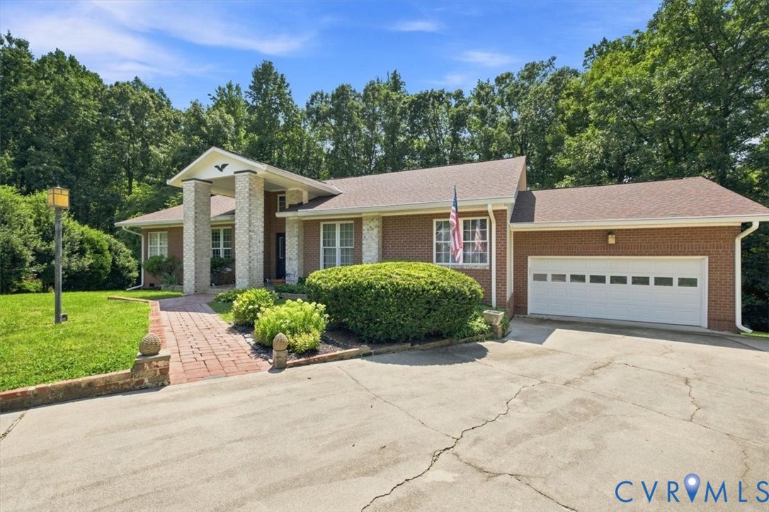 Modern brick residence featuring a two-car attached garage, circular driveway with brick pavers, mature landscaping with manicured hedges, and a contemporary architectural design with stone and brick exterior elements set on a generous lawn lot surrounded by established trees