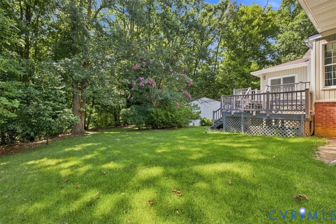 Expansive backyard featuring a manicured lawn, mature tree canopy providing natural shade, and a brick exterior home with an elevated composite deck and lattice railing