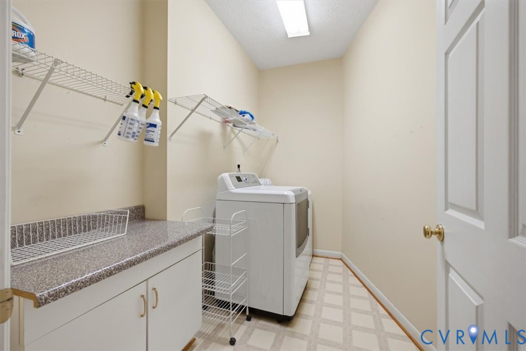 Dedicated laundry room featuring ample cabinetry with brushed gold hardware, speckled quartz countertop workspace, wall-mounted shelving for storage, ceramic tile flooring, and bright overhead lighting