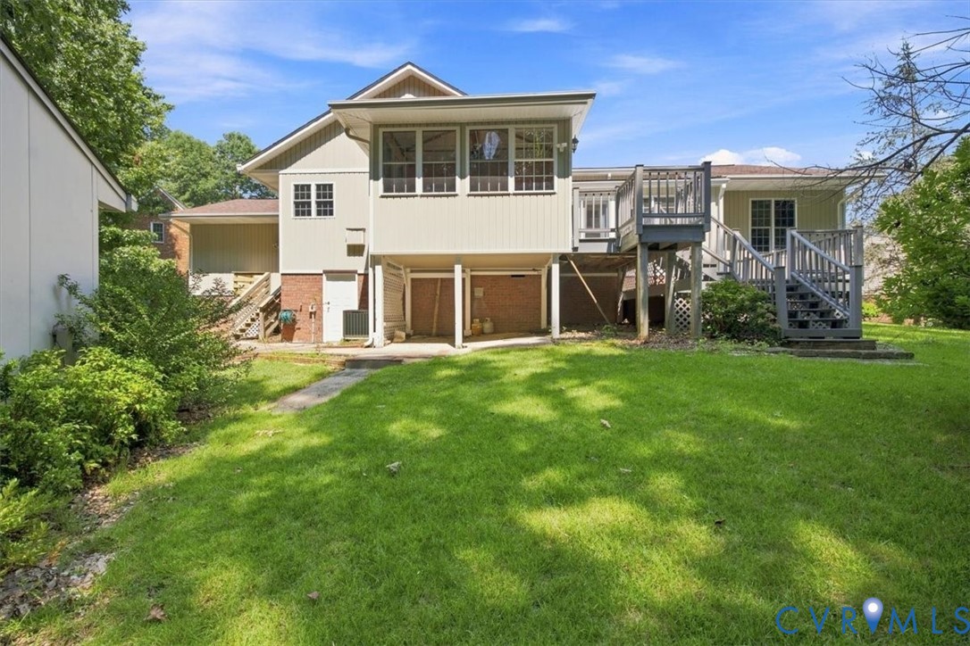 Spacious rear yard with mature landscaping and lush lawn, anchored by a two-story residence featuring brick and siding exterior, elevated deck with railings, and attached garage