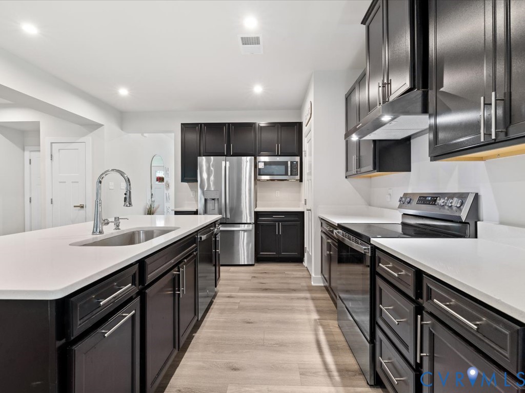Modern kitchen featuring a generous island with white quartz countertop and waterfall edge, stainless steel appliances including a built-in microwave, dark cabinetry with brushed metal hardware, white subway tile backsplash, and wood-finish flooring throughout the open layout