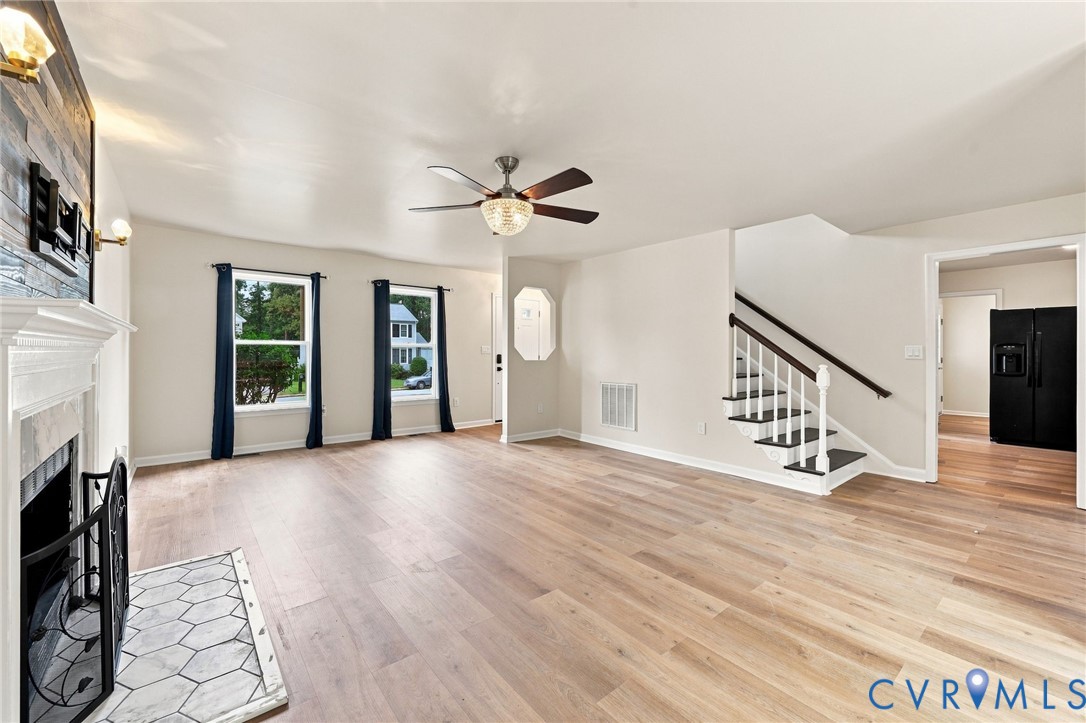 Light wood-finish flooring, a ceiling fan with integrated lighting, a fireplace with a white mantel and hexagonal tile hearth, and a staircase with white risers and a dark wood handrail