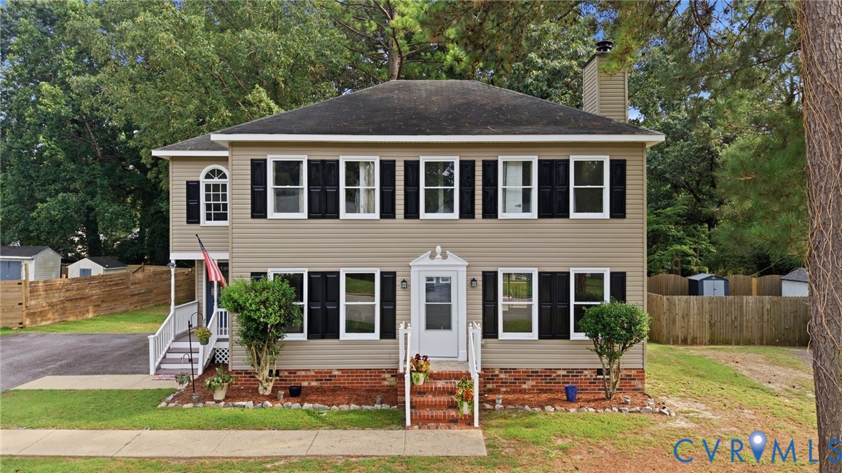 Two-story residence featuring light-toned siding, dark shutters, a front entry with brick-clad steps, and a side entrance with white railings Two-story residence featuring light-toned siding, dark shutters, a front entry with brick-clad steps, and a side entrance with white railings