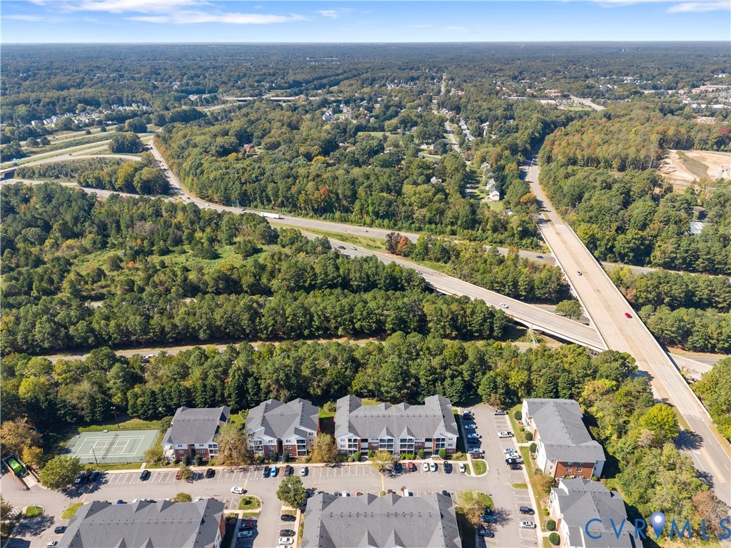 Aerial perspective of a multi-unit residential complex featuring gabled roofs, asphalt parking surfaces, a fenced tennis court, and surrounding mature woodland