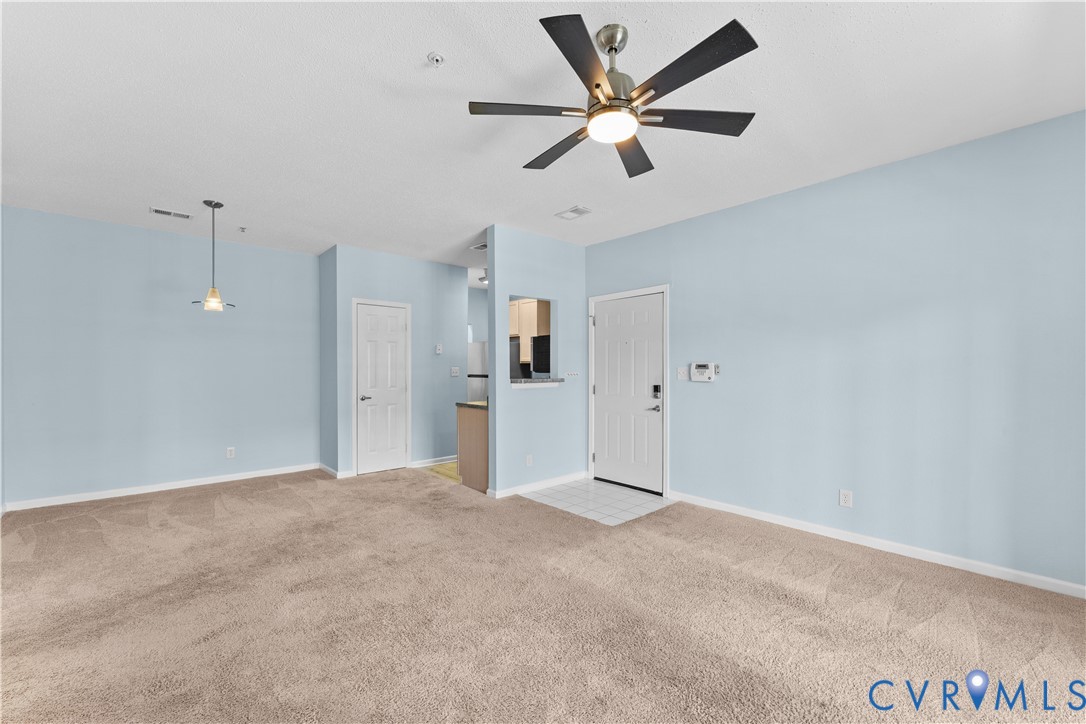 Living area featuring neutral carpeting, a central multi-blade ceiling fan with integrated lighting, a pendant fixture, and a pass-through opening to the kitchen