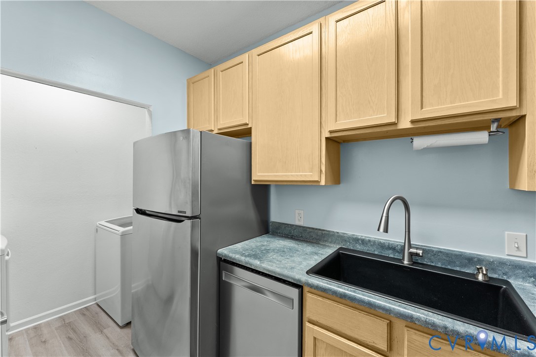 Utility space featuring light wood-finish cabinetry, stainless steel refrigerator and dishwasher, laminate countertop, and deep-basin sink with arched faucet