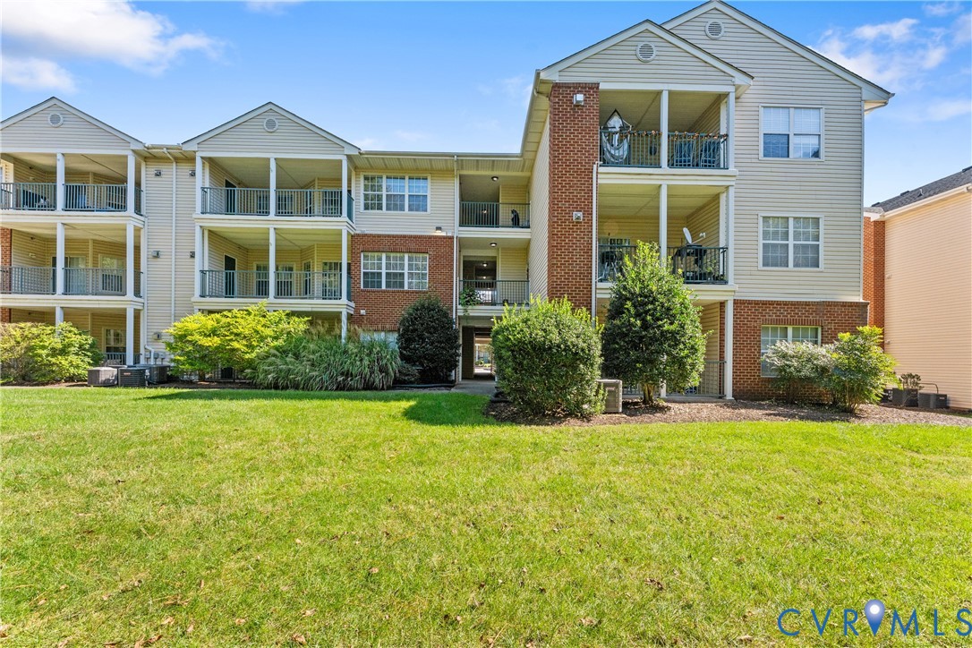 Multi-story building exterior featuring vinyl siding, brick masonry detailing, private covered balconies with metal railings, and an expansive lawn