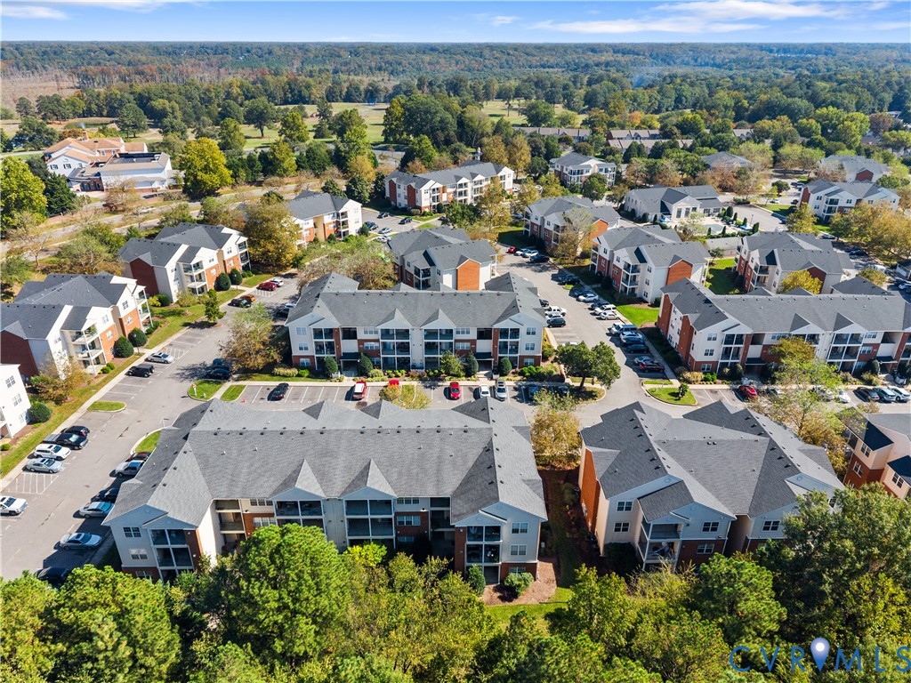 Aerial perspective of a multi-building residential complex featuring shingled roofs, private balconies, paved surface parking, and extensive mature tree canopy