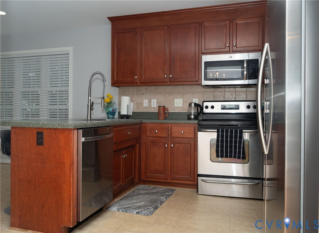 Kitchen featuring wood cabinetry, stainless steel appliances, a tiled backsplash, and an island with a built-in sink Kitchen featuring wood cabinetry, stainless steel appliances, a tiled backsplash, and an island with a built-in sink