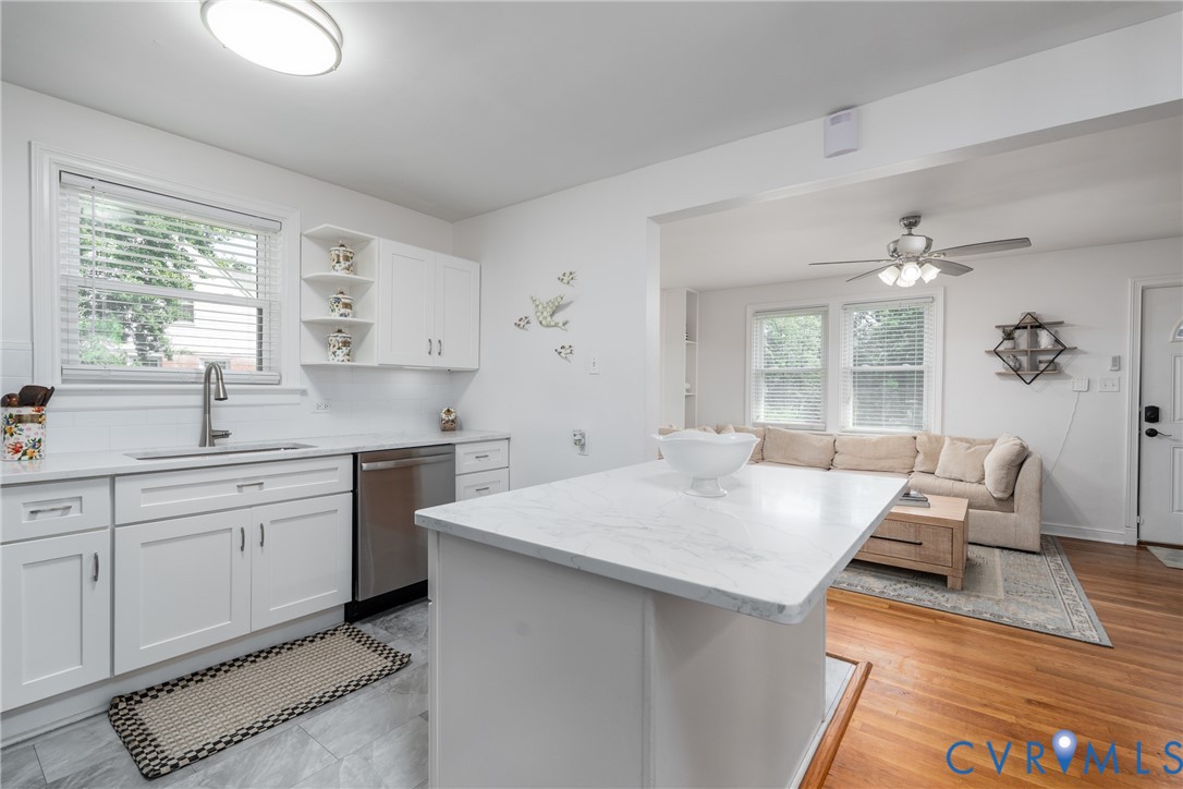 White kitchen cabinetry, undermount sink with gooseneck faucet, light-toned countertop, stainless steel dishwasher, and wood-finish flooring in the adjacent room