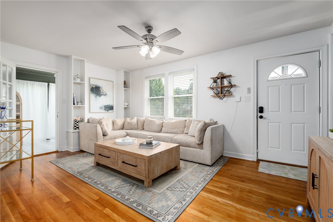 Hardwood flooring, ceiling fan with integrated lighting, built-in shelving, windows with blinds, and a paneled entry door with transom