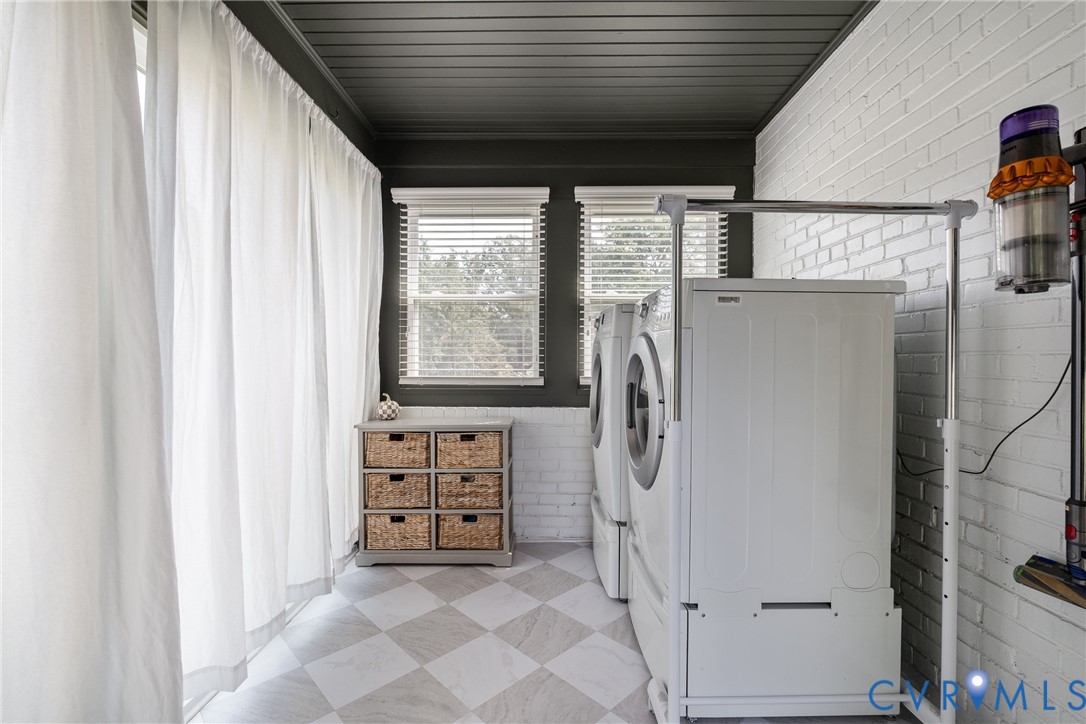 White brick accent wall, checkered flooring, dark paneled ceiling, and two windows with blinds