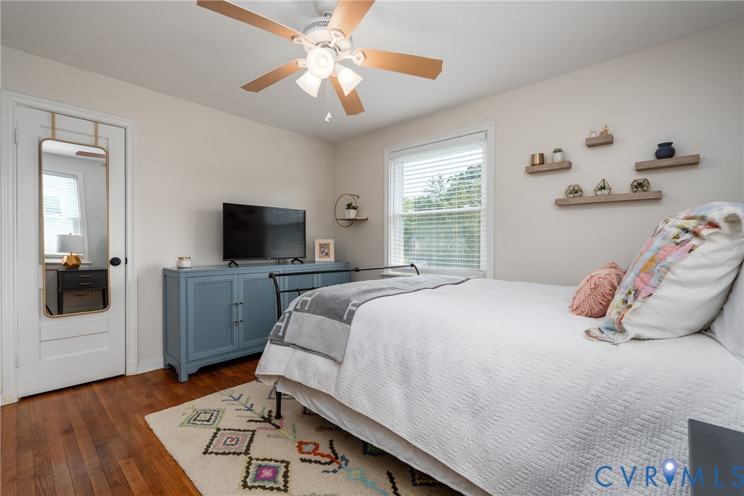 Hardwood flooring, ceiling fan with light fixture, large window with blinds, and built-in wall shelving