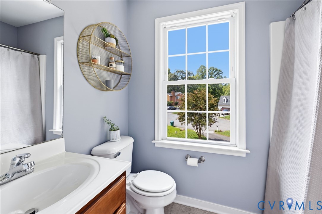 White single-hung window with grid, wood-finish vanity with countertop sink, polished chrome faucet, white toilet, and tile flooring