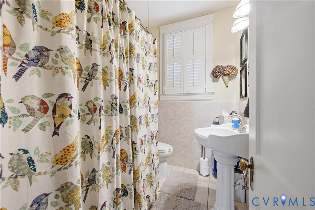 Main floor bath with white pedestal sink, chrome fixtures, tile flooring, window with white louvered shutters, and wall-mounted lighting