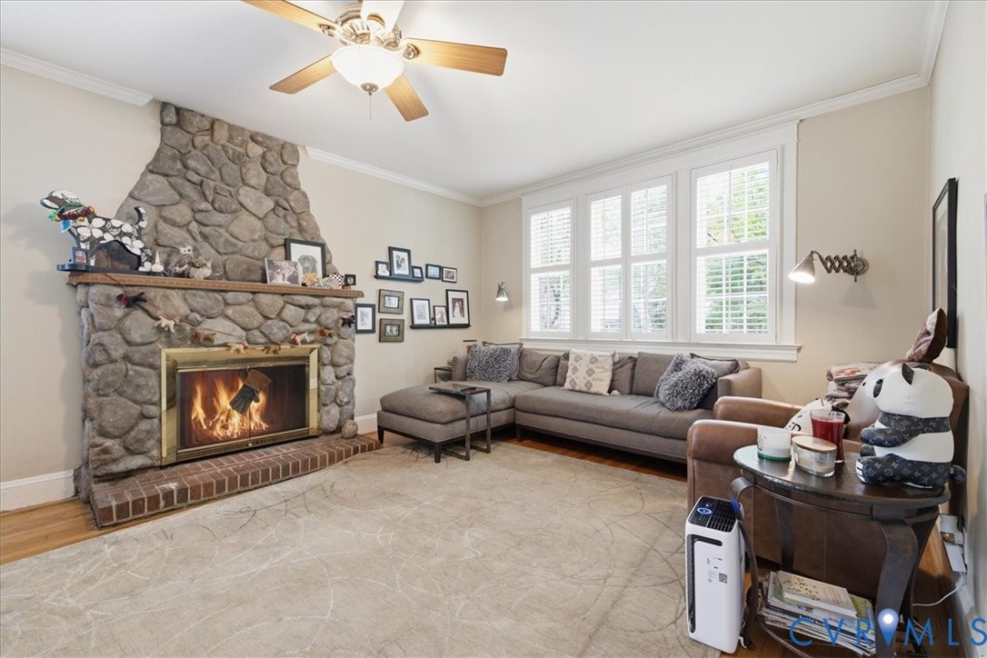 Stone look-clad fireplace with brick hearth, multiple transom windows with plantation shutters, crown molding, and a ceiling fan with integrated lighting Stone look-clad fireplace with brick hearth, multiple transom windows with plantation shutters, crown molding, and a ceiling fan with integrated lighting