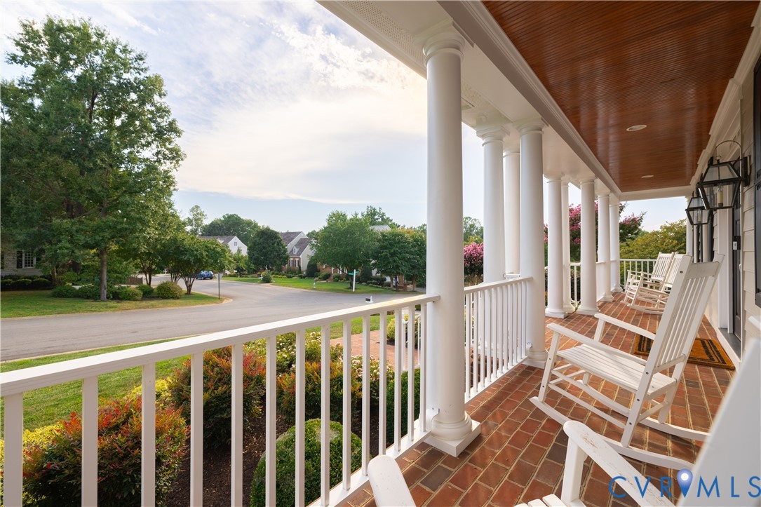 Expansive front porch featuring brick flooring, white columns, wood-finish ceiling, and exterior wall lanterns