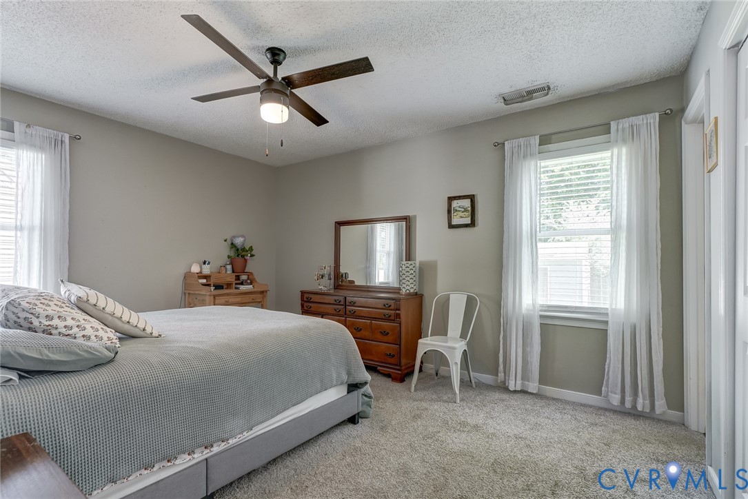 Plush carpet flooring, neutral painted walls, ceiling fan with integrated lighting, and multiple windows with blinds