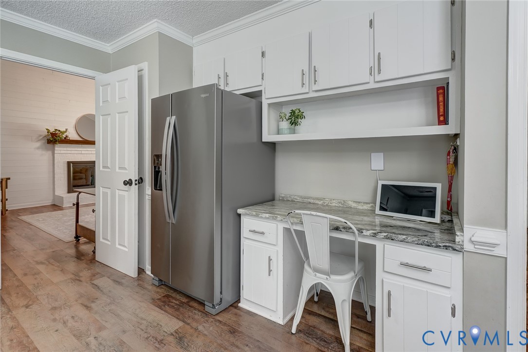 Wood-finish flooring, built-in desk with stone-finish countertop, white cabinetry with silver-finish hardware, stainless steel refrigerator, and crown molding