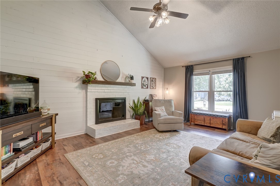 Vaulted ceiling with a ceiling fan, white shiplap accent wall, brick fireplace with a wood-finish mantel, and wood-finish flooring