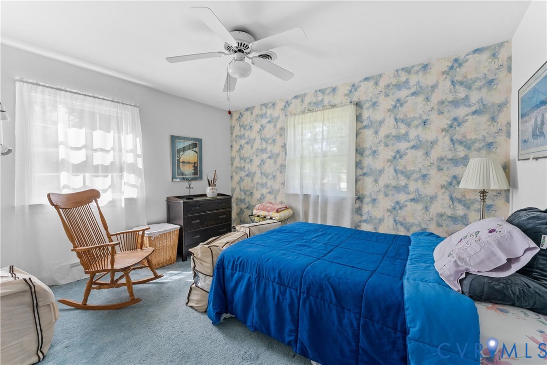 Textured wall covering accent, ceiling fan with integrated lighting, two windows with sheer curtains, light-colored carpeting