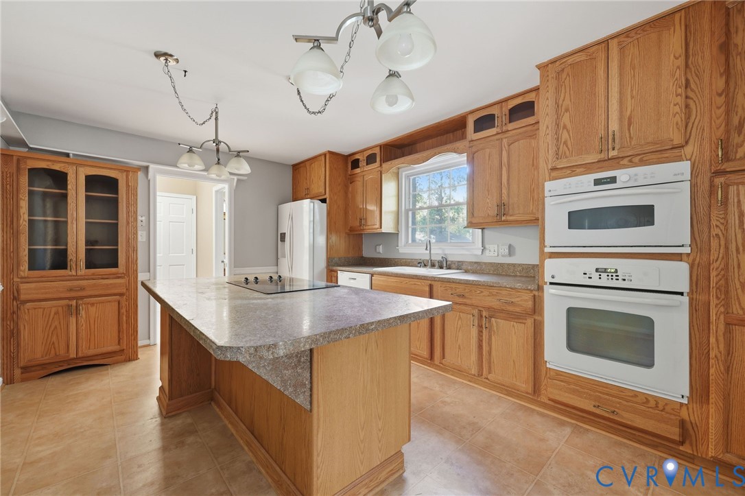 Kitchen featuring oak cabinetry, stone-finish countertops, a central island with a cooktop, double wall ovens, and tile flooring
