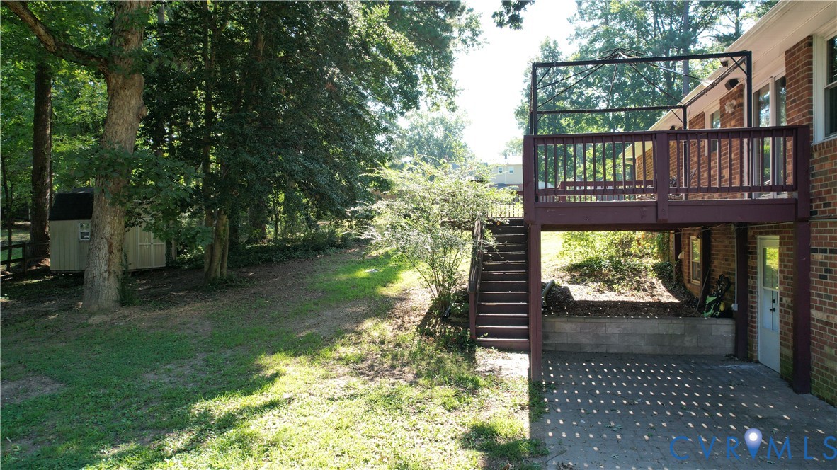 Elevated wood deck with railings, exterior brick wall, paved patio area, and mature trees