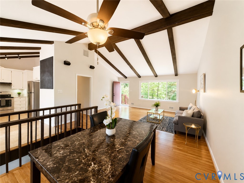 Open-concept living space featuring exposed wood beam ceiling with integrated ceiling fan, expansive windows with natural light, warm wood-finish flooring, dark stone countertop island, and black metal railing detail