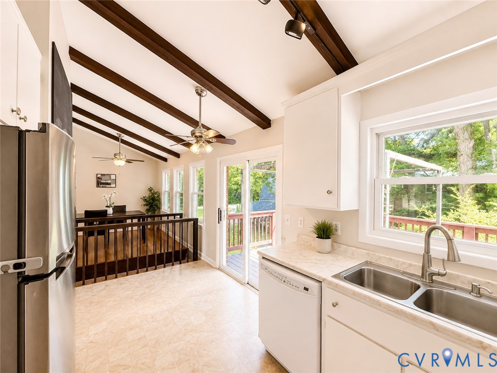 Vaulted ceilings with exposed wood beams, stainless steel appliances, white cabinetry, double basin sink, and a sliding glass door to an outdoor area