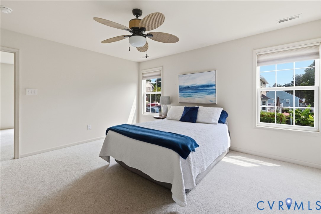 Light-filled room featuring neutral wall tones, plush carpeting, a ceiling fan with woven blades, and multiple windows