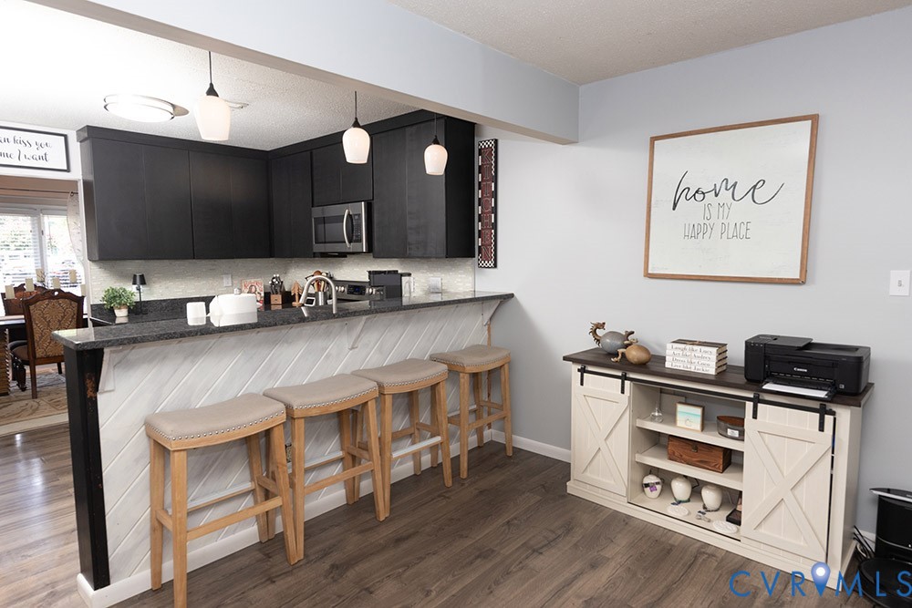 Modern kitchen and entertaining space featuring black cabinetry with stainless steel appliances, quartz waterfall island countertop with seating for three, pendant lighting, and wood-finish flooring throughout