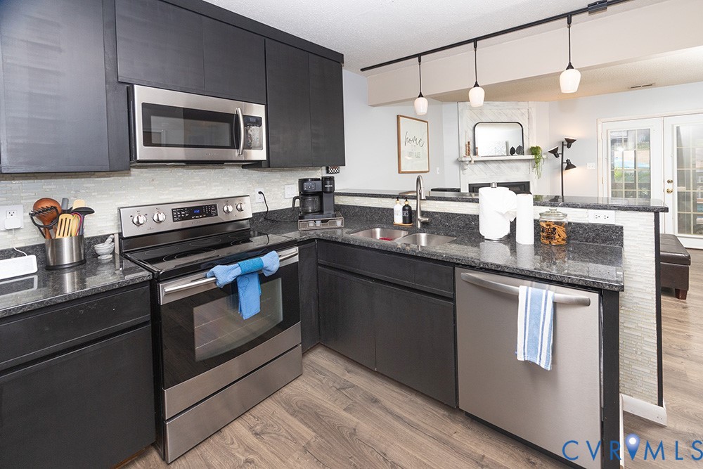 Kitchen featuring dark cabinetry, stainless steel appliances, speckled stone countertops, a tile backsplash, and wood-finish flooring