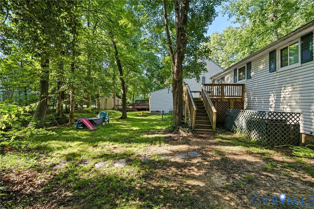 Wood deck with railing and stairs, siding exterior, multiple windows with shutters, mature trees, and a grassy lawn