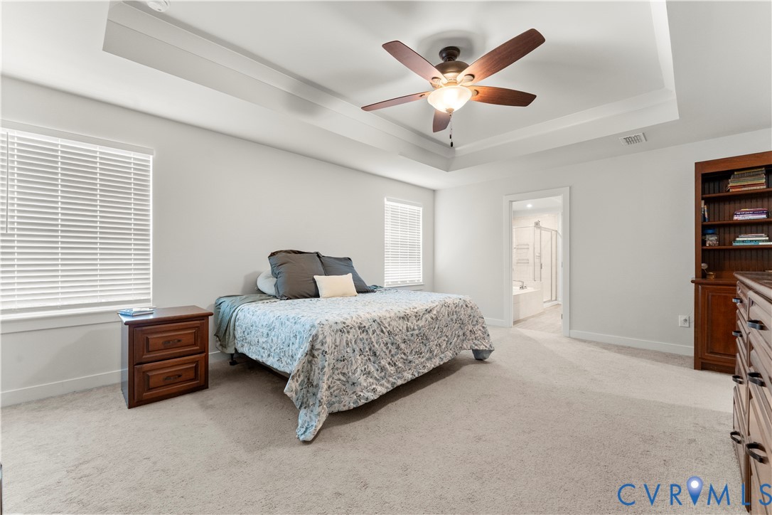 Tray ceiling, ceiling fan with light fixture, light-colored carpeting, white window blinds, and built-in shelving