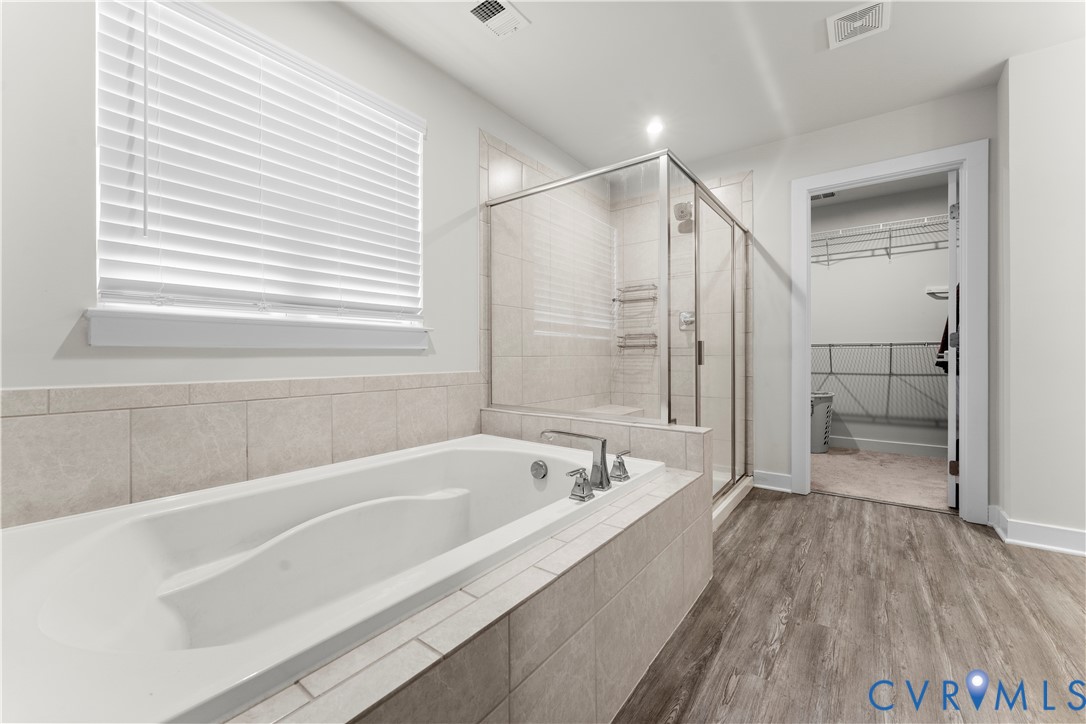 Soaking tub with tile surround, glass-enclosed shower, wood-finish flooring, and a window with blinds