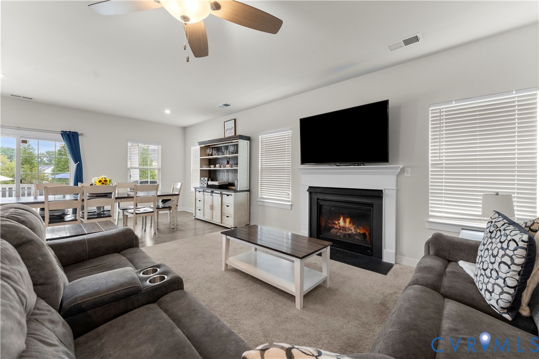 Open-concept living space featuring a fireplace with a white mantel, ceiling fan, carpeted and wood-finish flooring, and white window blinds