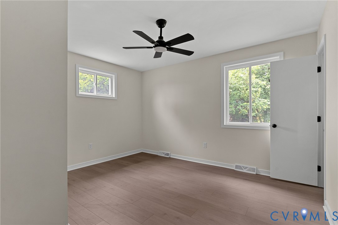 Neutral-toned room featuring wood-finish flooring, a modern black ceiling fan with lighting, white trim, and two windows providing natural illumination