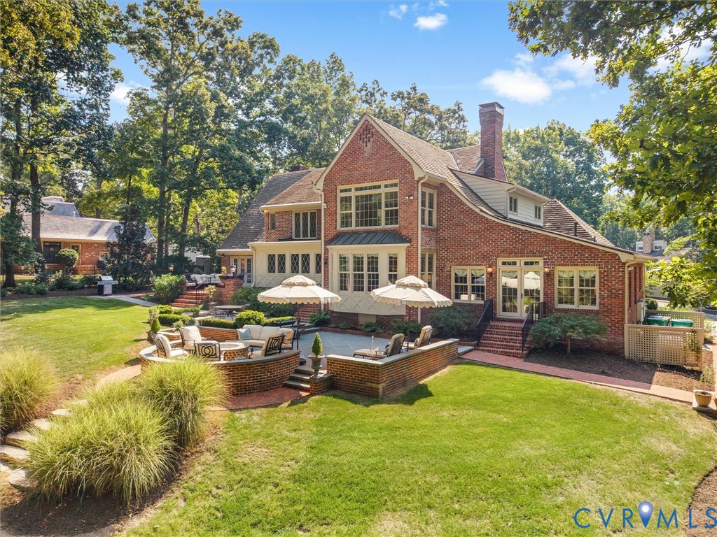 Brick exterior with multiple dormer windows, a tiered hardscape patio, an expansive lawn, and mature trees