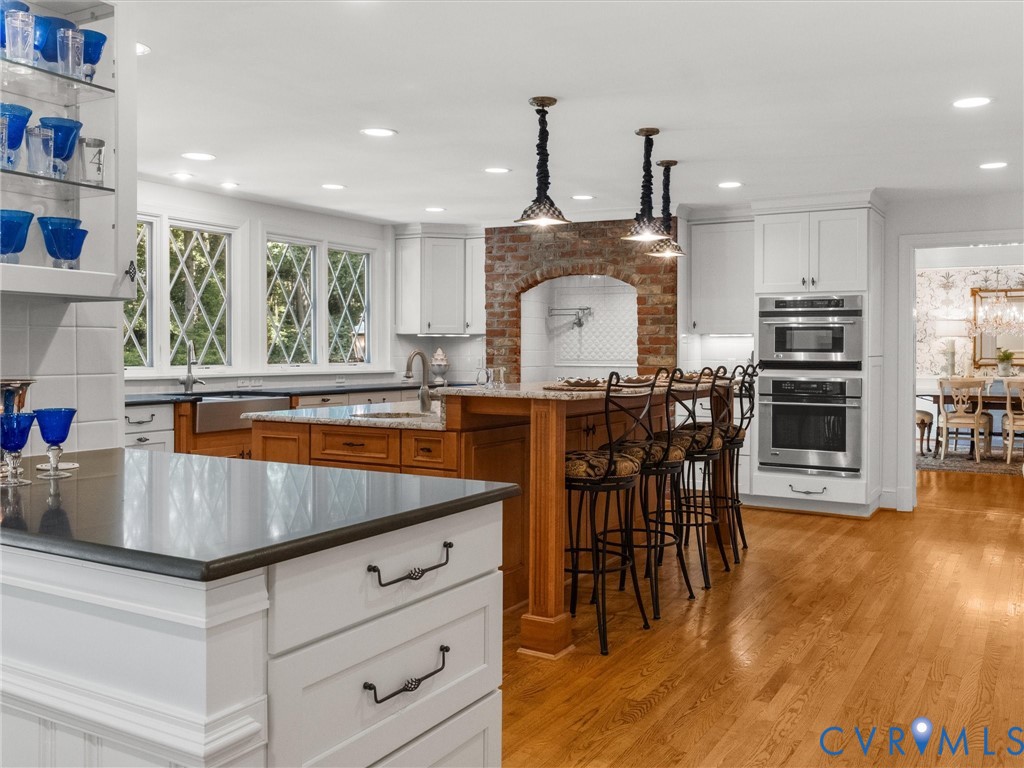 Wood-finish flooring, white cabinetry, a brick archway with a tiled backsplash, built-in stainless steel appliances, and an island with a breakfast bar