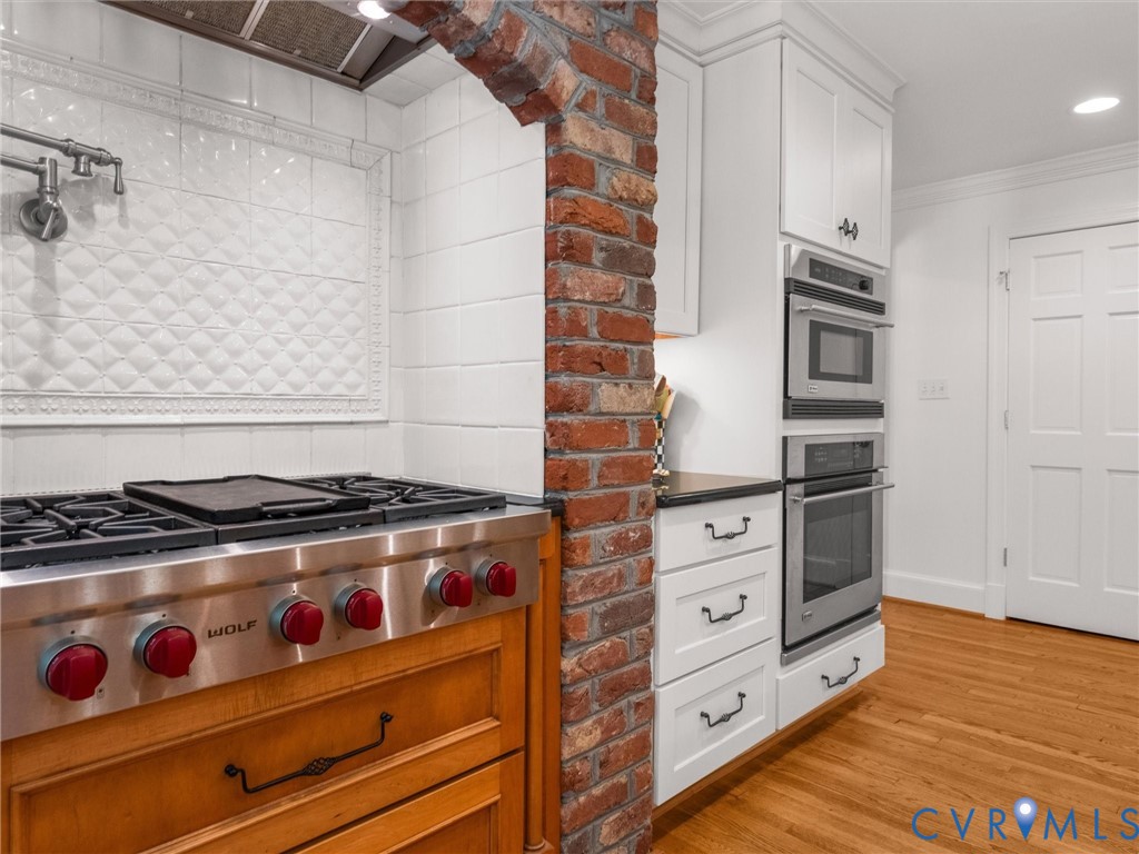 Stainless steel range with red knob accents, brick archway, white cabinetry, integrated stainless steel microwave and oven, and wood-finish flooring