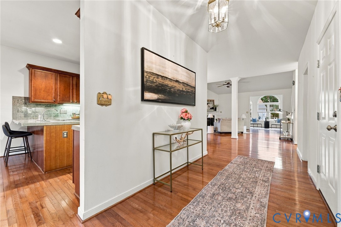 Hardwood-finish flooring, vaulted ceiling with chandelier, arched doorway, recessed lighting, and kitchen area with wood cabinetry and stone-finish backsplash