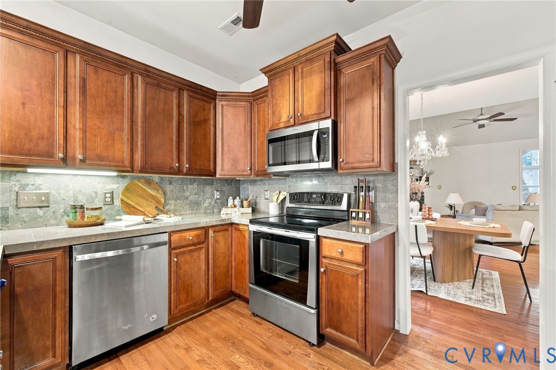 Wood-finish cabinetry with crown molding, stainless steel appliances, natural stone backsplash, and light-toned countertops